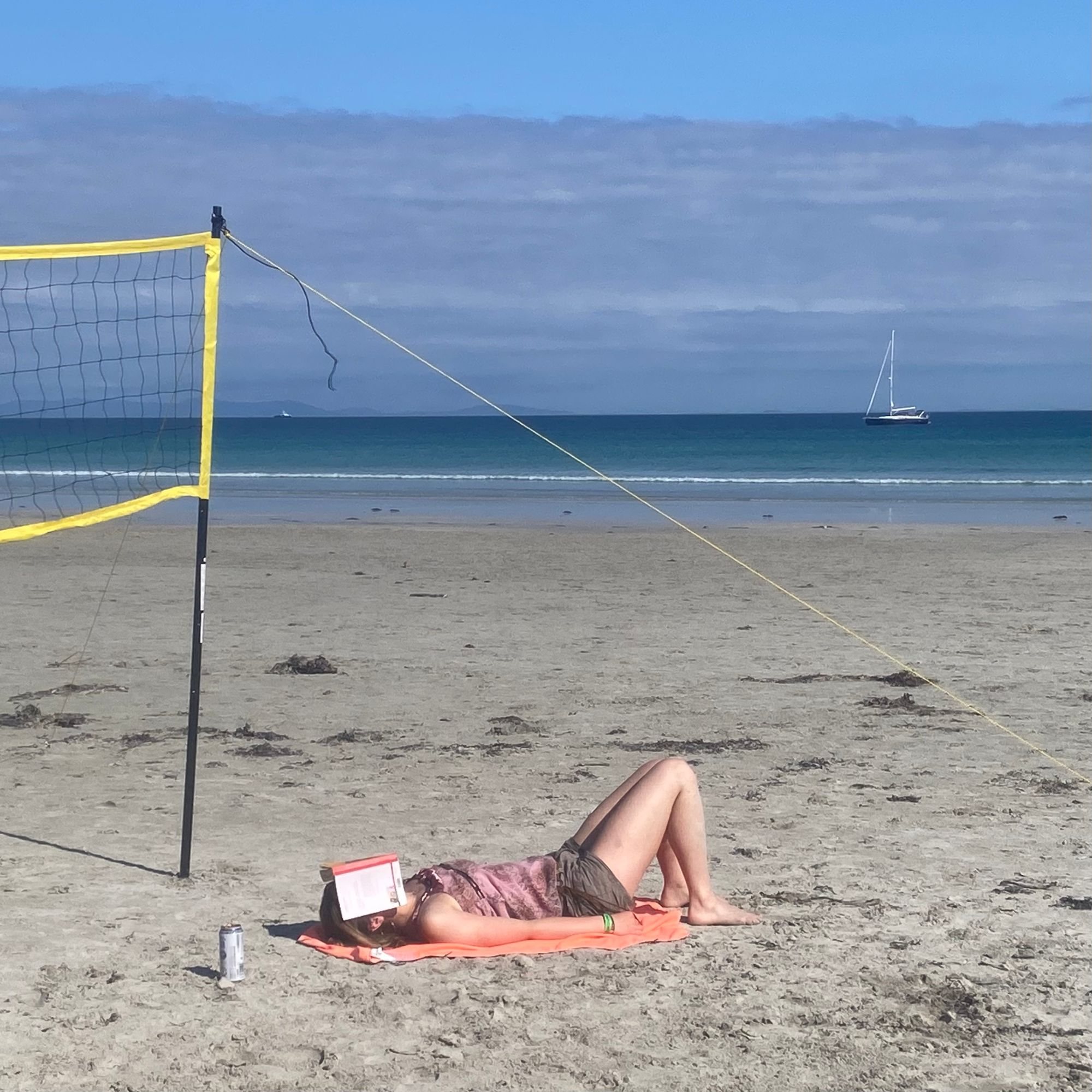 Photograph by Tom Speedy. A woman lies asleep on a sandy beach beside a yellow volleyball net, an open paperback resting over her face, a drinks can by her head. Blue sea and a moored yacht behind.