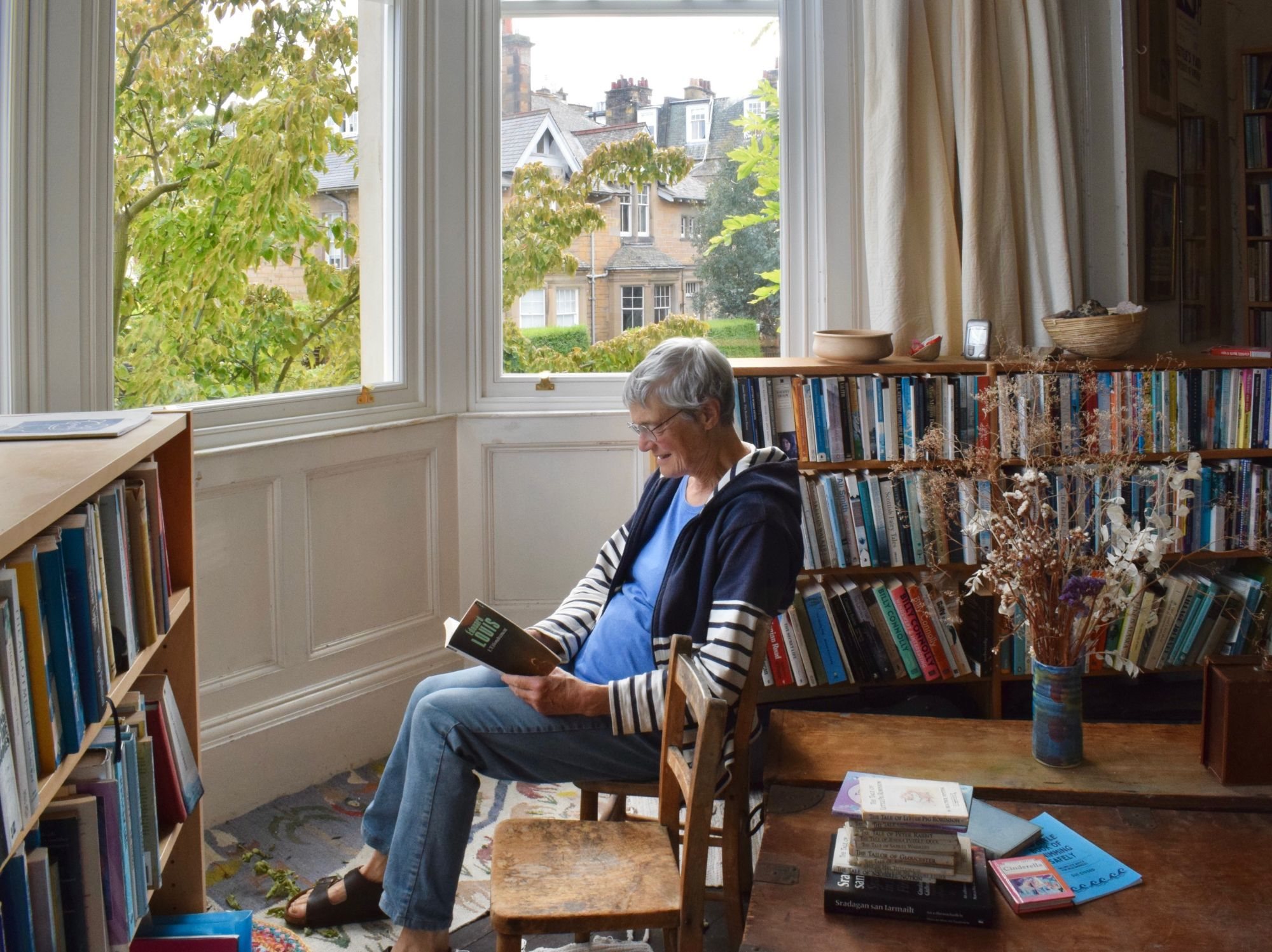 Photograph by Shakya Seresinghe. A grey-haired woman in a striped cardigan sits on a wooden chair reading a paperback in a bay window, surrounded by full bookshelves, with sandstone houses and trees outside.