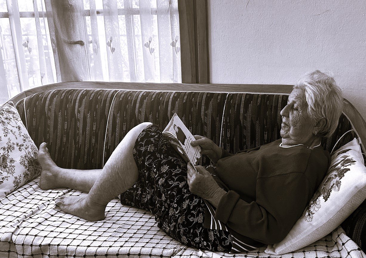 Afternoon Reading, by Zeynep Özge Özdemir Diler. Black-and-white photograph of an elderly woman lying back on a patterned sofa by a curtained window, bare feet stretched out, reading a children's picture book.