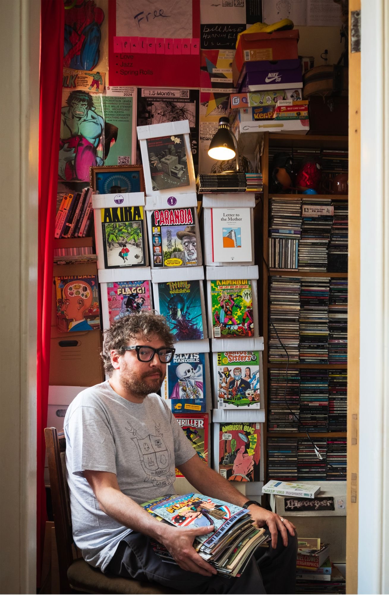 Photograph by Alejandro Basterrechea. A man with curly hair and thick-framed glasses sits holding a stack of comics, looking at the camera, in front of a wall of boxed comics, posters and shelves of CDs.