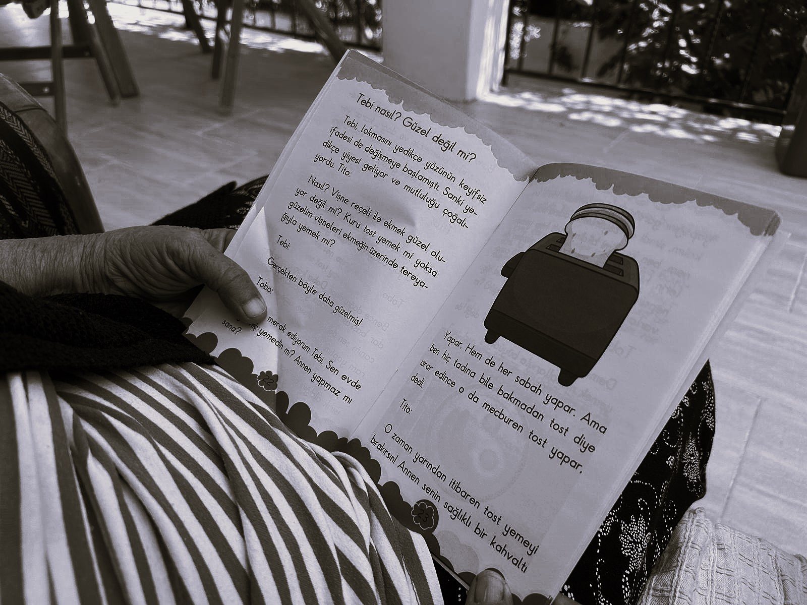 The Hands That Read, by Zeynep Özge Özdemir Diler. Black-and-white close-up of an elderly woman's hand holding open a Turkish children's book on her striped lap.