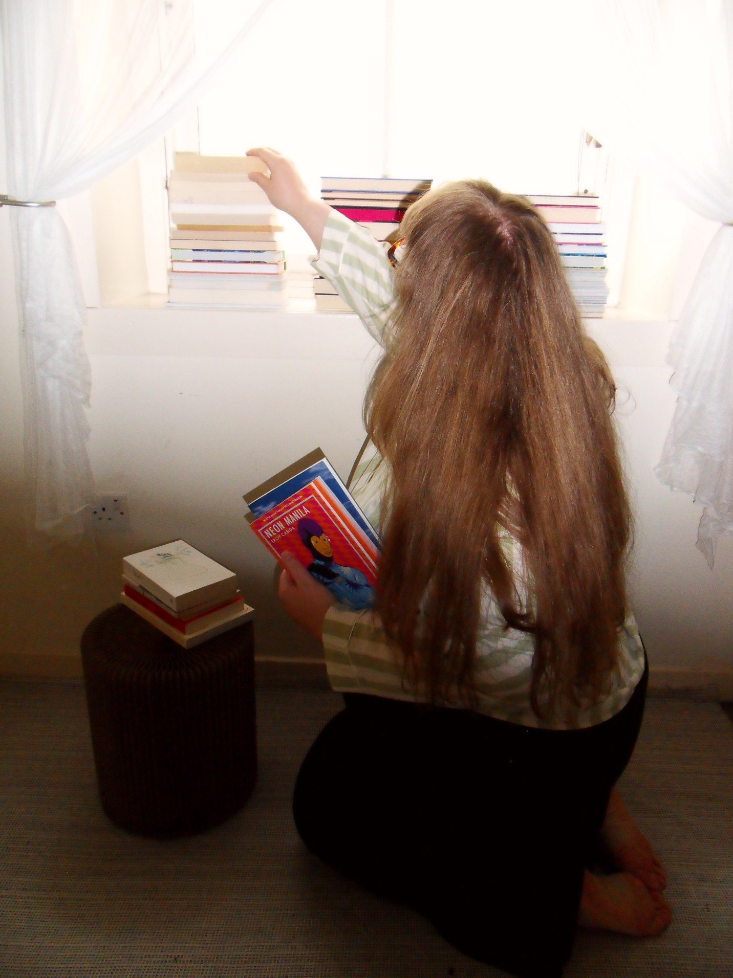 Photograph by Alexandra Teleková. A woman with long hair kneels on the floor, seen from behind, stacking books with their spines facing a bright window, holding more books in her other hand.