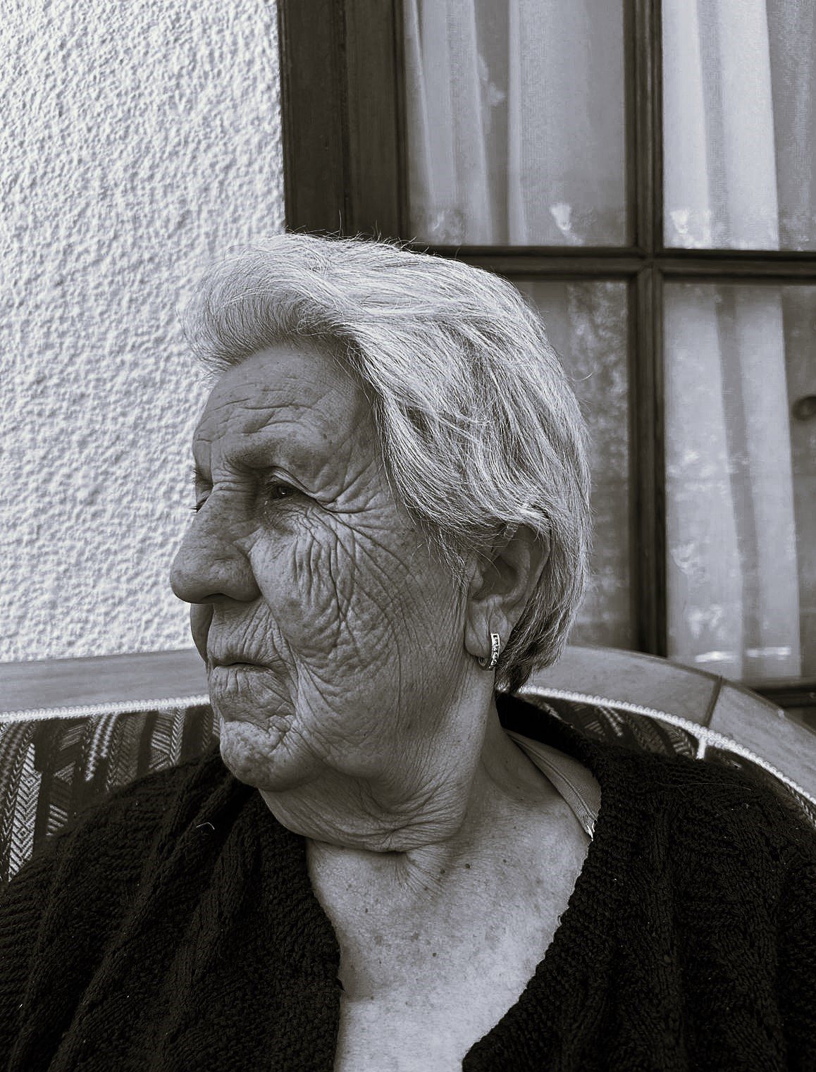 Photograph by Zeynep Özge Özdemir Diler. Black-and-white portrait of an elderly woman in profile, with short grey hair and a small earring, seated by a curtained window.
