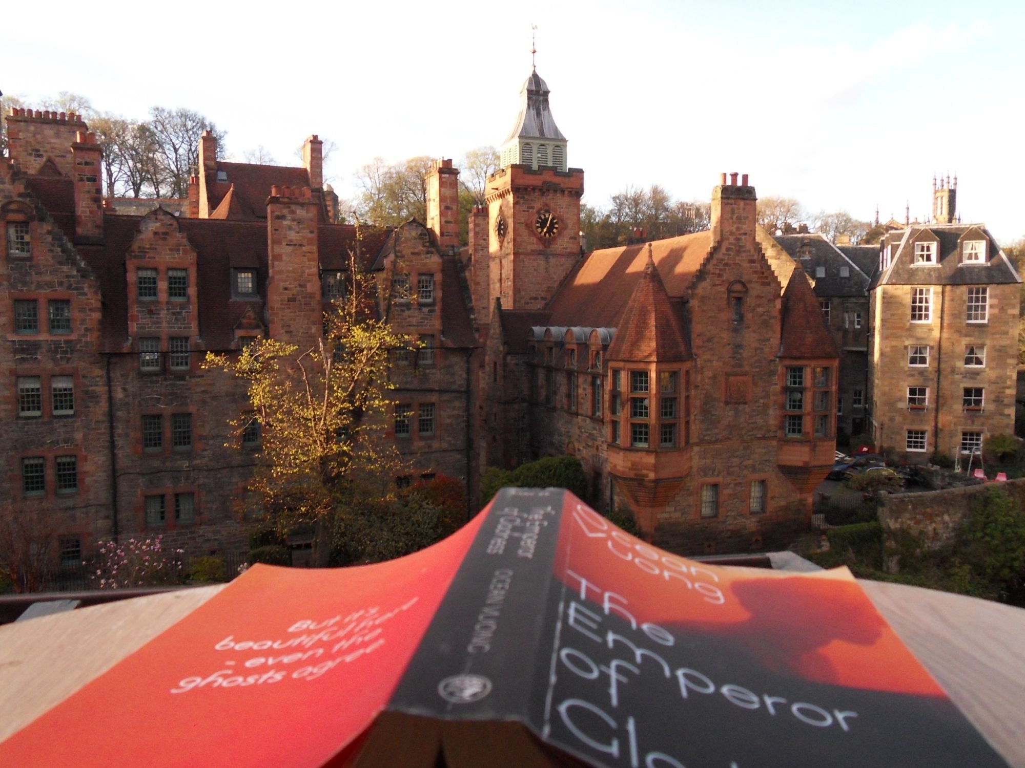 Photograph by Alexandra Teleková. An open paperback, The Emperor of Gladness by Ocean Vuong, lies face down on a windowsill in the foreground. Beyond it, the red sandstone buildings and clock tower of Well Court in Dean Village catch the evening light.