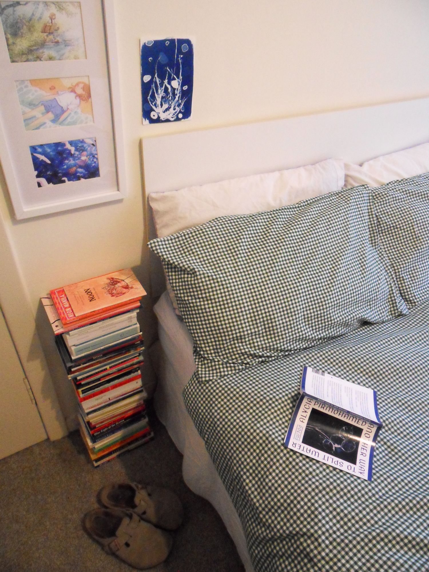 Photograph by Alexandra Teleková. A tall pile of books beside a bed with green gingham covers, an open poetry book on the bed, slippers on the floor and a blue cyanotype and framed illustrations on the wall above.
