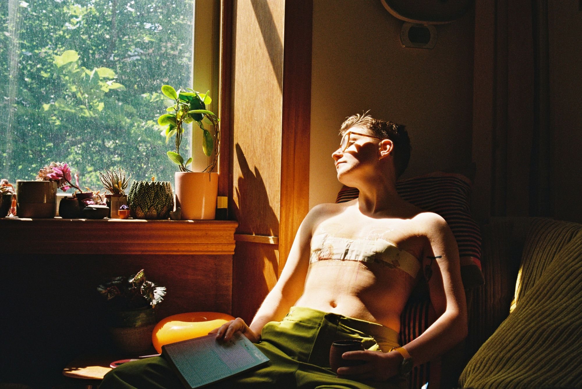 room to breathe, by Elsa van der Wal. Resting in the intermittent sun by a window, eyes closed and face turned to the light, a book open on the lap between naps. Houseplants line the windowsill.