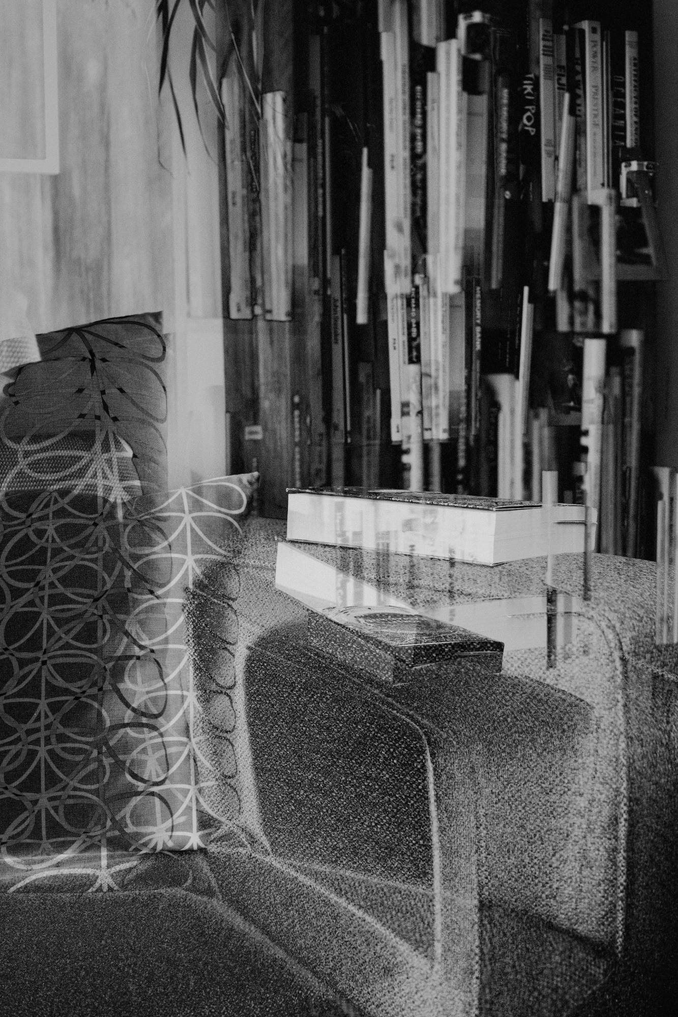 Photograph by Josh Murfitt. Black-and-white multiple exposure of the corner of a sofa with patterned cushions and books on the armrest, bookshelves behind.