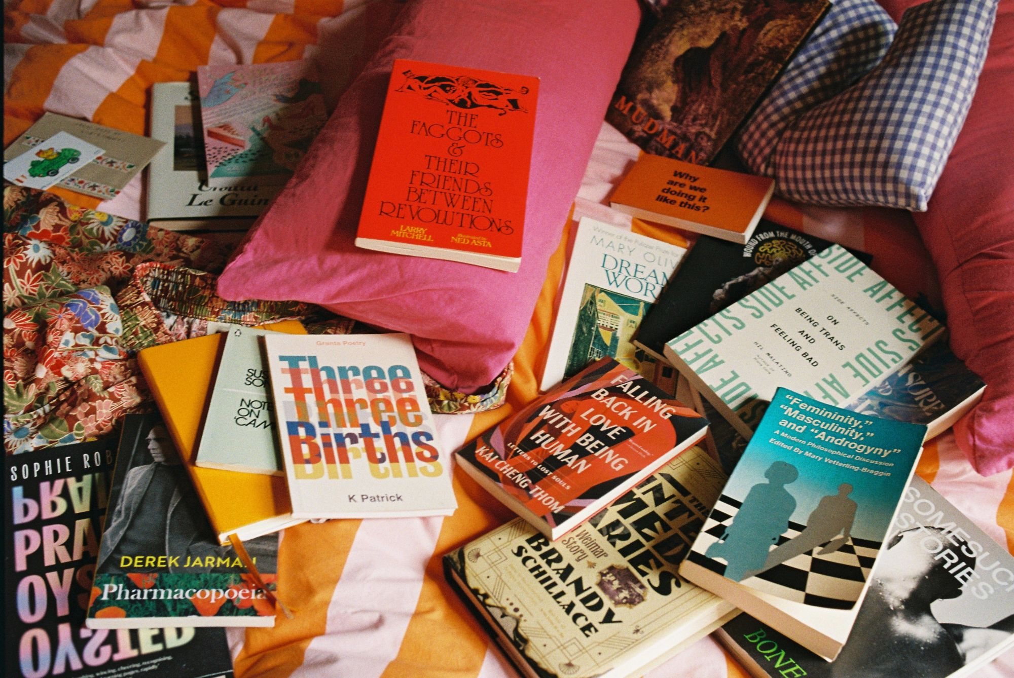Photograph by Elsa van der Wal. A still life of books spread across a bed with orange and pink striped sheets, among pillows and floral pyjamas.