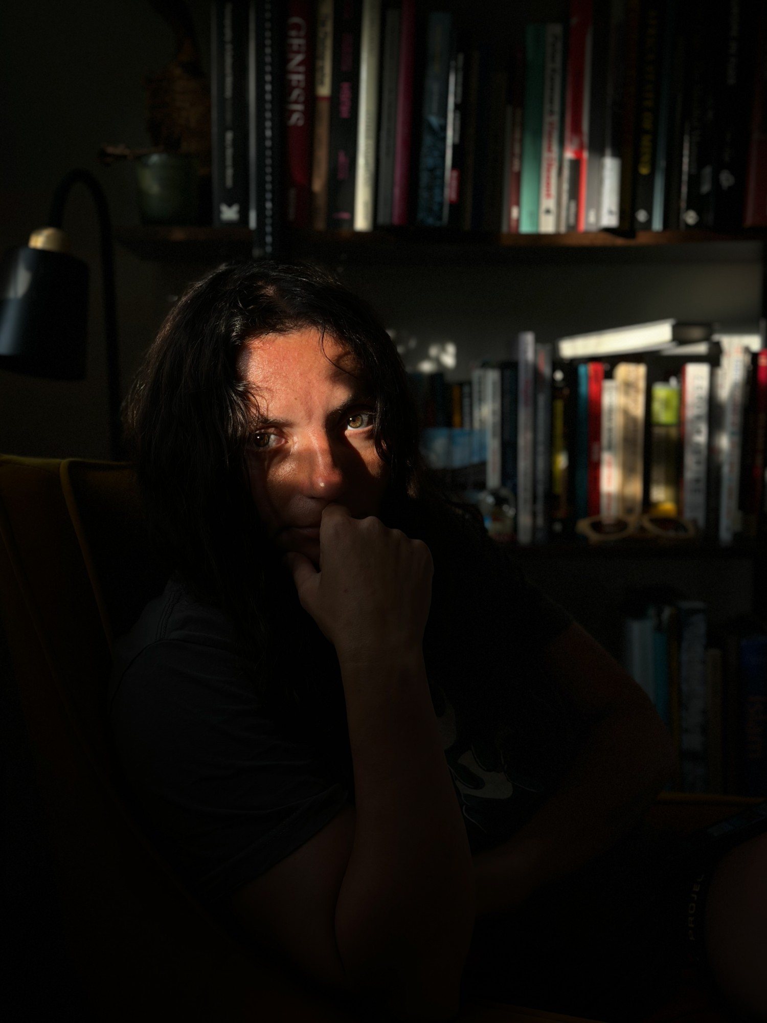 Photograph by Matt Aslett. Portrait of a woman with long dark hair in a dark room, a band of sunlight falling across her eyes, her chin resting on her hand, bookshelves behind her.