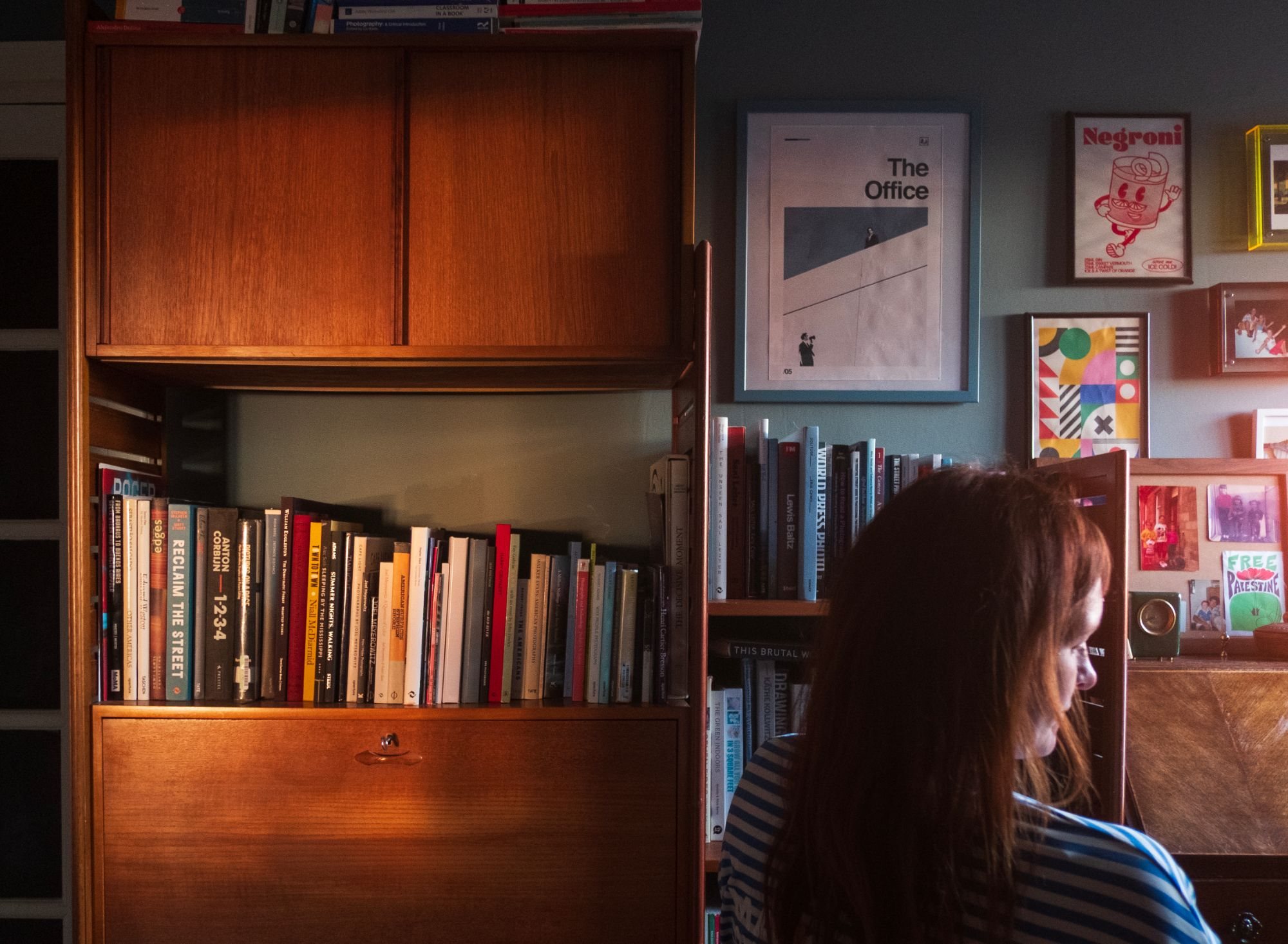 Simone, by Alejandro Basterrechea. A woman with long red hair, seen from behind and in profile, sits beside a teak bookcase of photography books lit by a shaft of sunlight, with framed posters on the wall beyond.