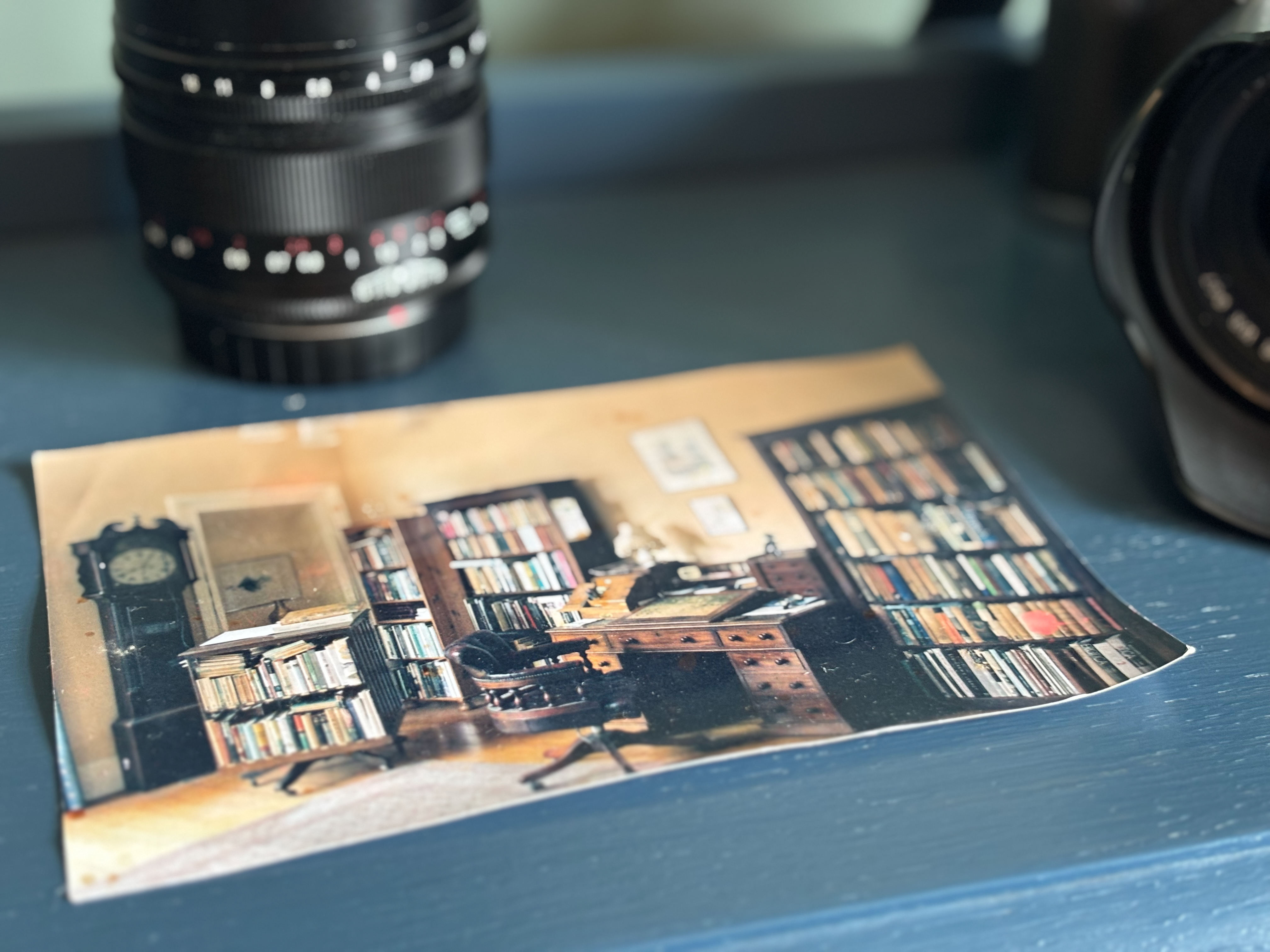 A printed photograph of a book-filled study resting on a desk beside camera lenses